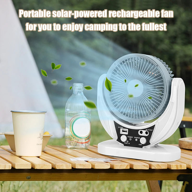 Portable solar-powered rechargeable fan on a camping table with a bottle and cup, surrounded by greenery.
